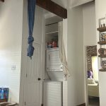 A narrow laundry nook with a stacked washer and dryer, a blue birth sling hanging from a ceiling beam, and a glimpse of a child's play area.