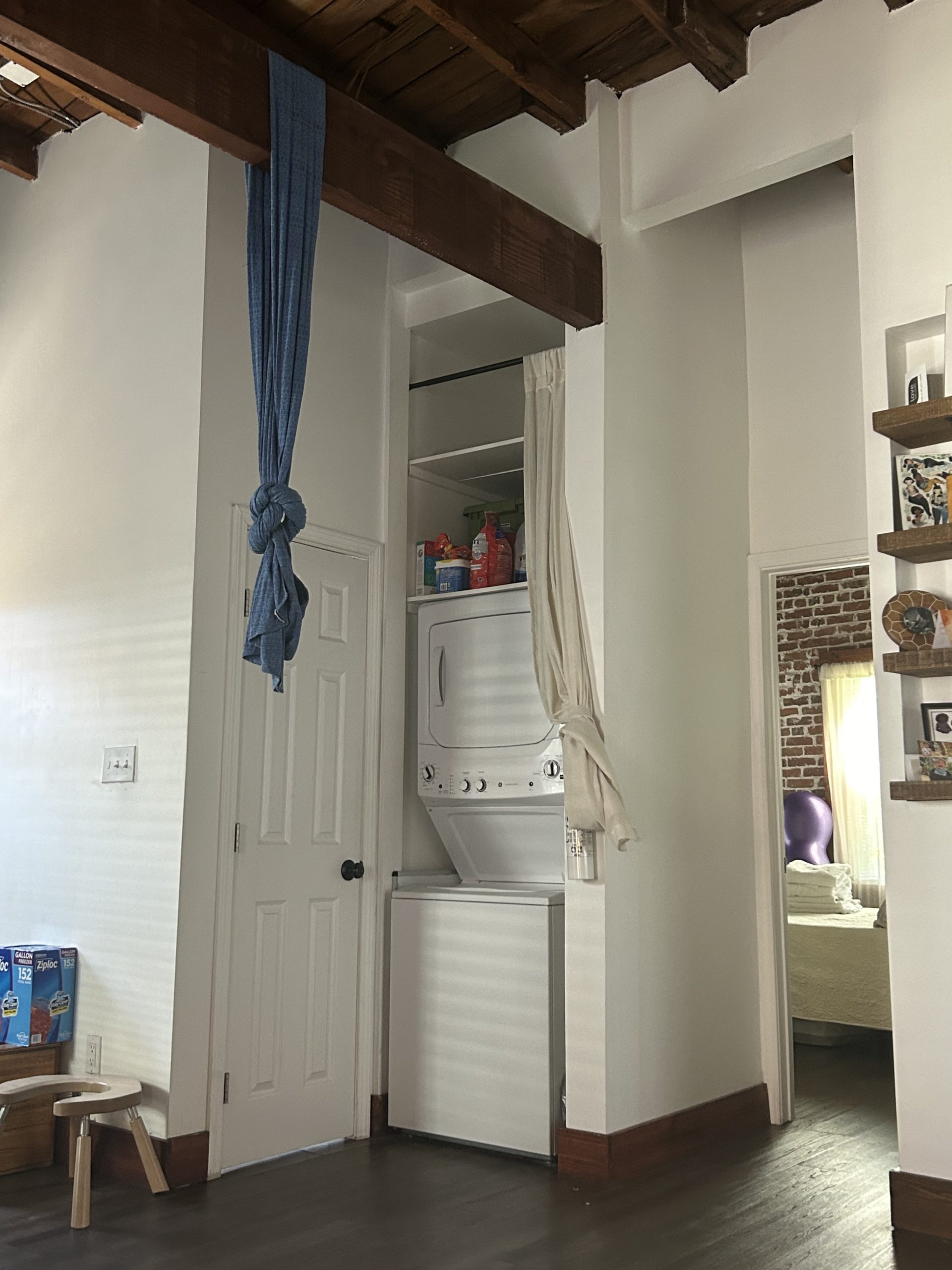 A narrow laundry nook with a stacked washer and dryer, a blue birth sling hanging from a ceiling beam, and a glimpse of a child's play area.