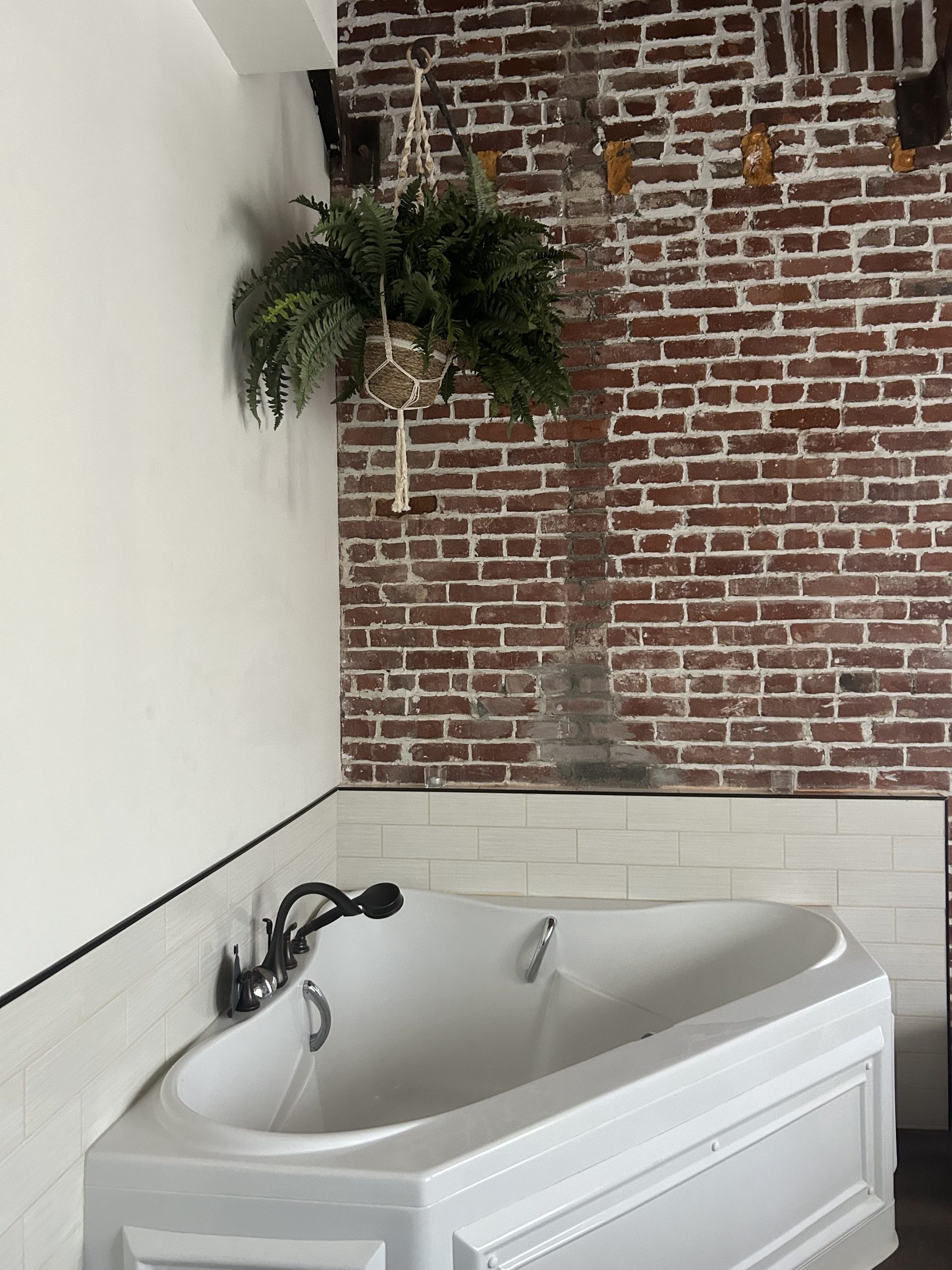 A soaking tub in a corner under a hanging plant and exposed red brick walls, creating a spa-like atmosphere.