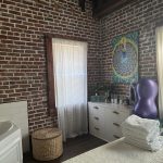 A cozy birthing room with brick walls, a massage chair, a cabinet with folded towels, and a radiant mandala wall hanging.