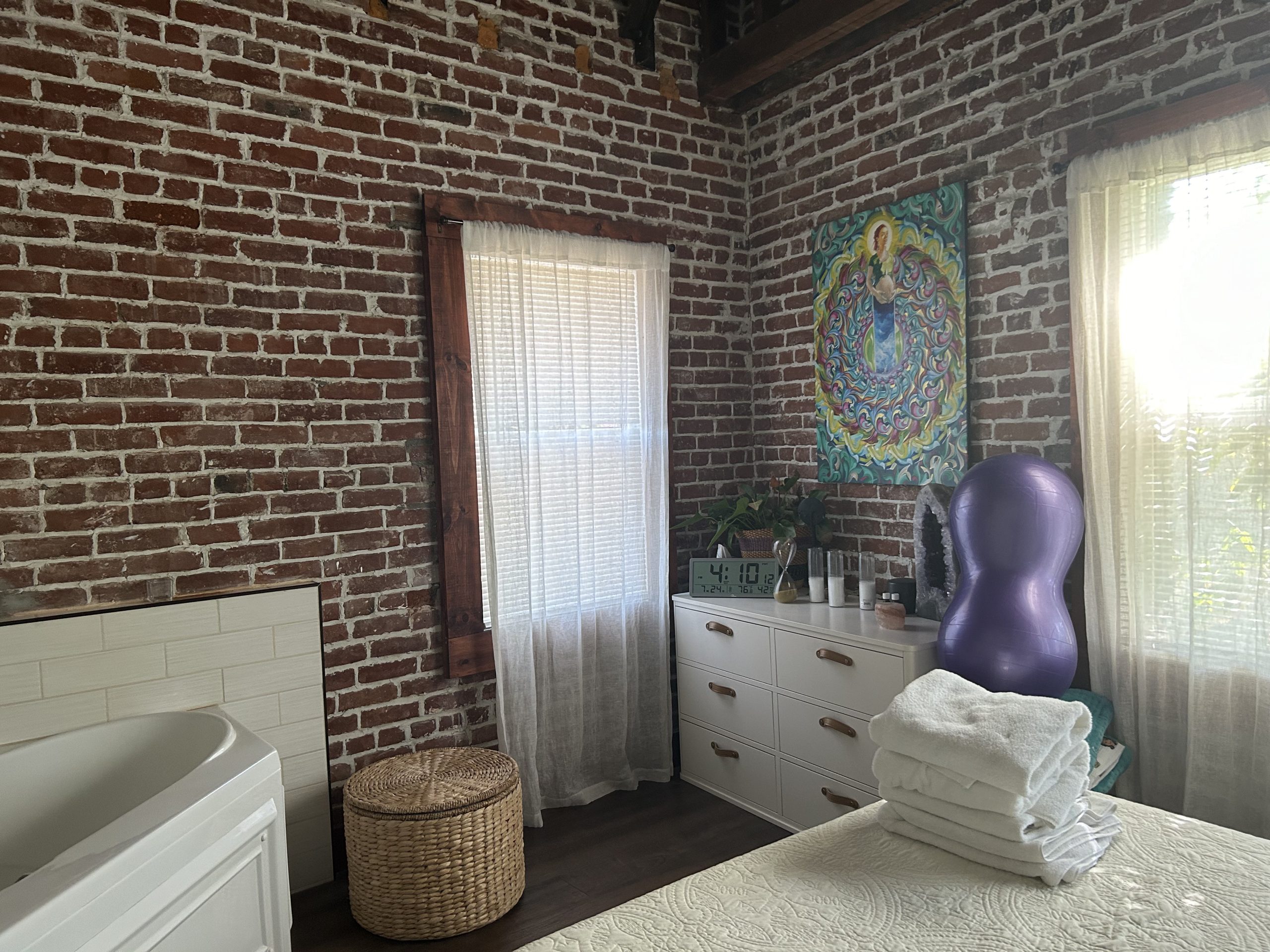 A cozy birthing room with brick walls, a massage chair, a cabinet with folded towels, and a radiant mandala wall hanging.