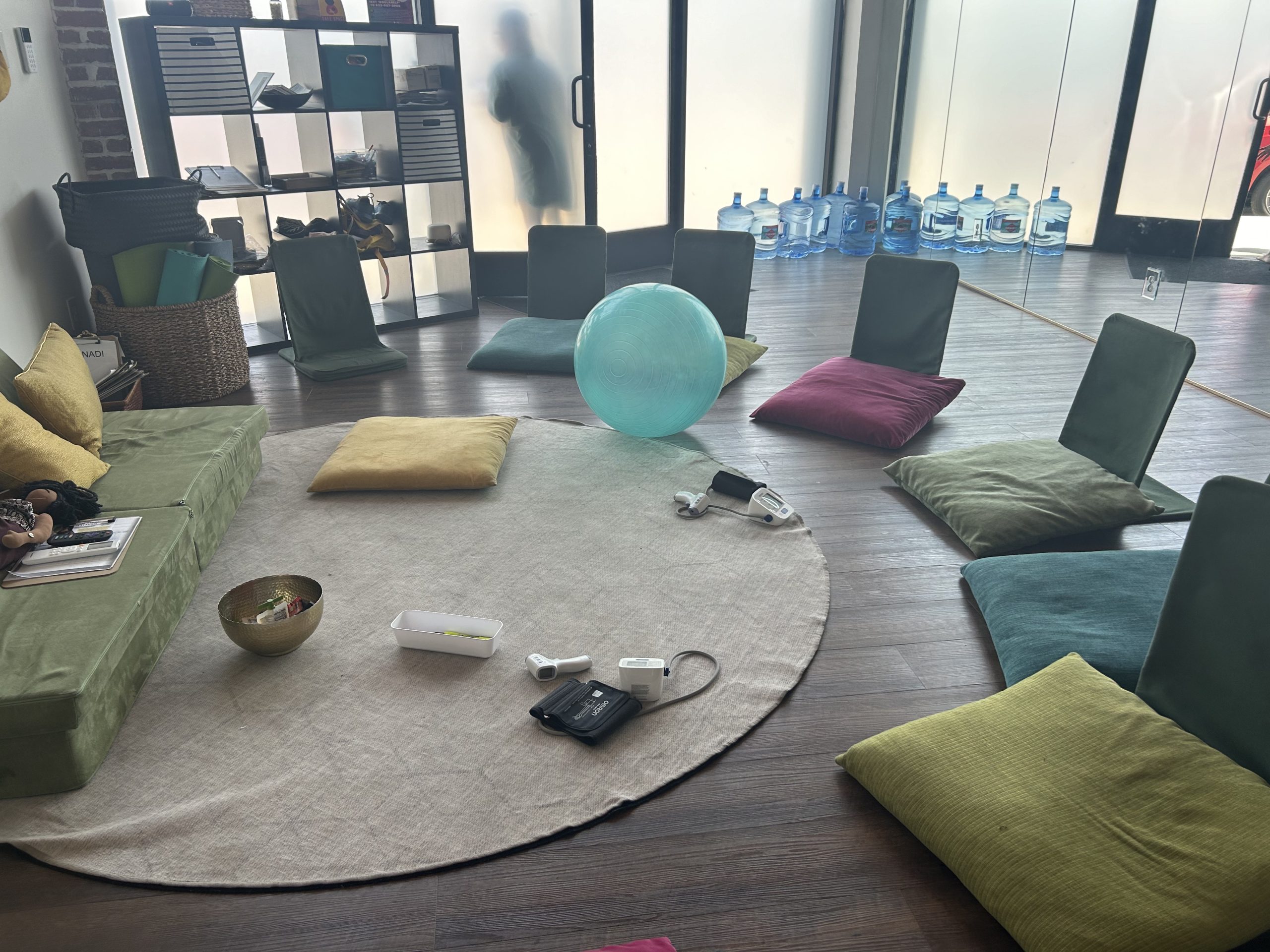 A lounge area with low green and gold floor cushions around a small table with candles and bowls, next to a wall of shelves and water bottles
