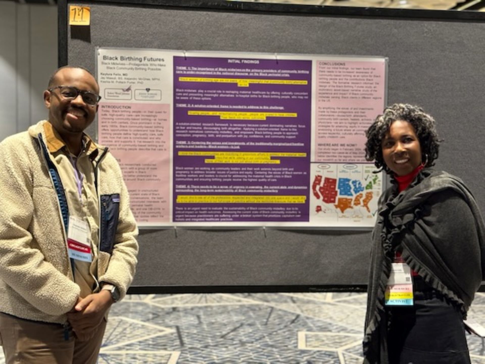 Two people standing and smiling in front of a research poster presentation featuring findings on a purple and yellow tri-panel academic display.